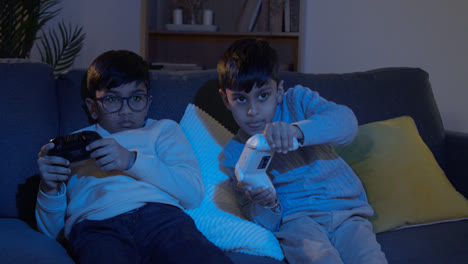 Two-Young-Boys-Sitting-On-Sofa-At-Home-Playing-With-Computer-Games-Console-On-TV-Holding-Controllers-Late-At-Night